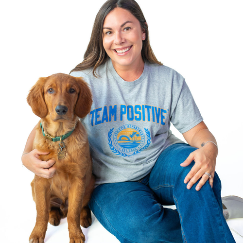 A smiling woman sitting on a white studio floor with a golden retriever dog. She is wearing a heather gray t-shirt with a blue graphic that reads 'TEAM POSITIVE' and a crest, paired with blue jeans.