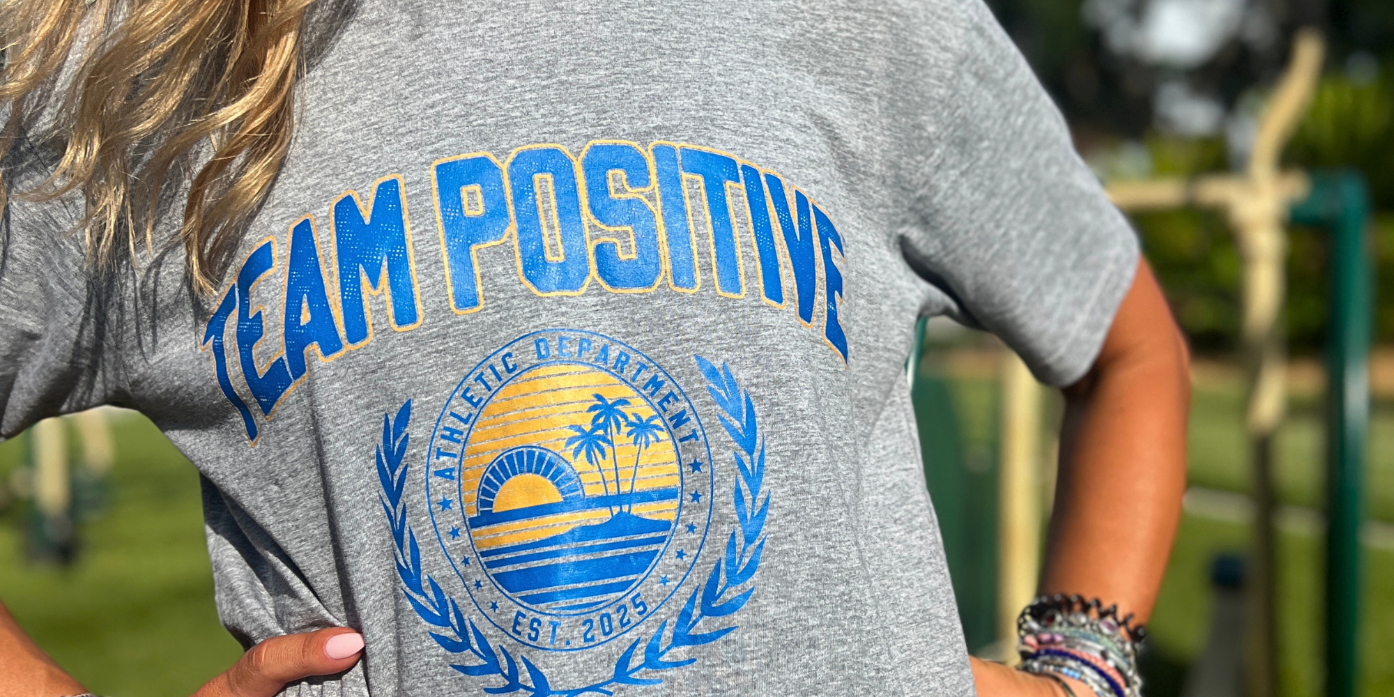 A close-up of a person wearing a heather gray t-shirt with a blue and yellow graphic that reads 'TEAM POSITIVE' and features a circular ocean sunset design.